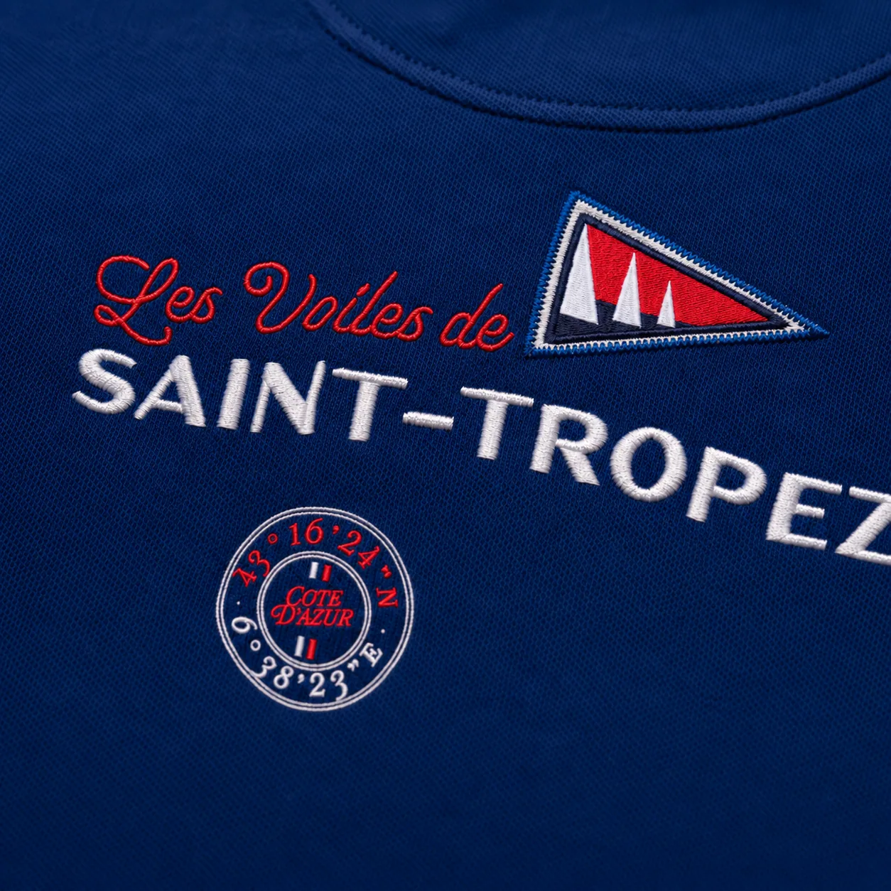 Macro de la broderie du dos, sous l'encolure : « Les Voiles de Saint-Tropez » et les coordonnées du golfe, sur maille piquée marine.