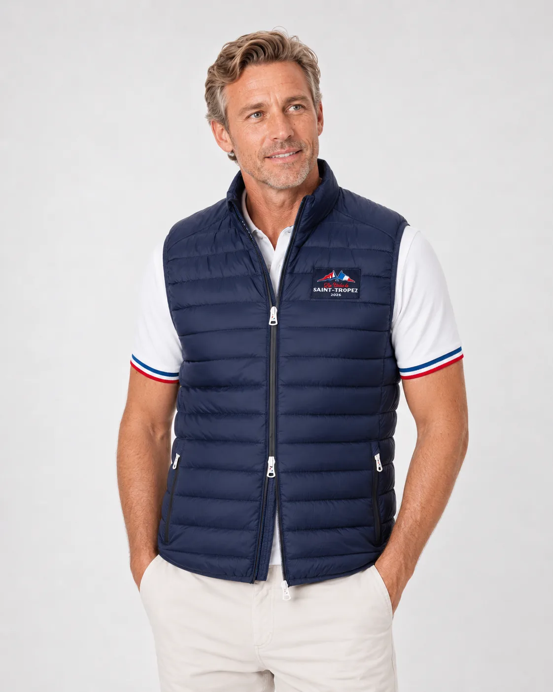 Doudoune Sans Manches 26 bleu marine portée sur un polo blanc : col montant, zip à deux curseurs, poches latérales zippées et étiquette tissée de l'édition sur la poitrine.