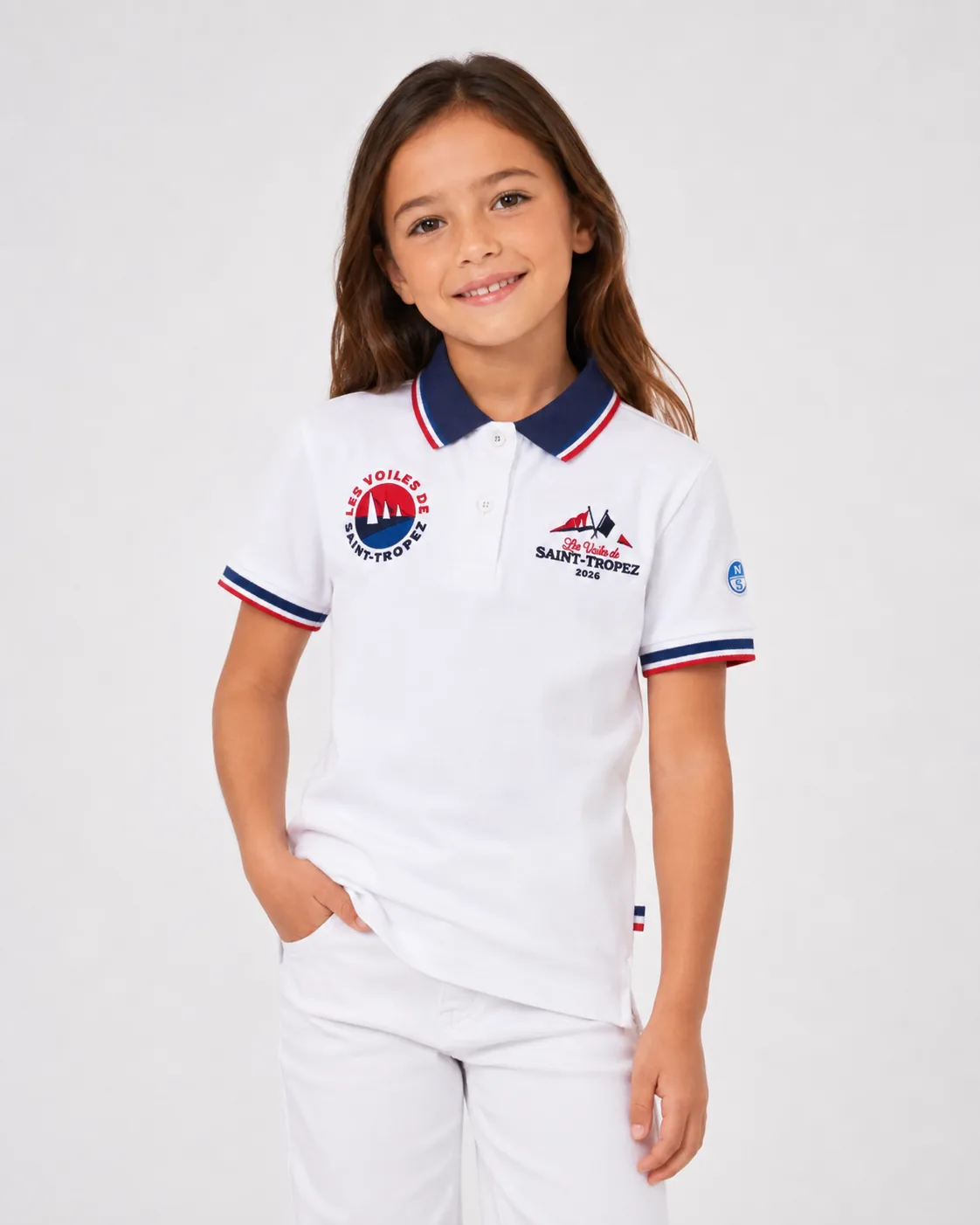Polo Kids 26 blanc porté de face par une enfant : col marine à galon tricolore, écusson rond à gauche, mention Les Voiles de Saint-Tropez 2026 à droite et écusson North Sails à la manche.