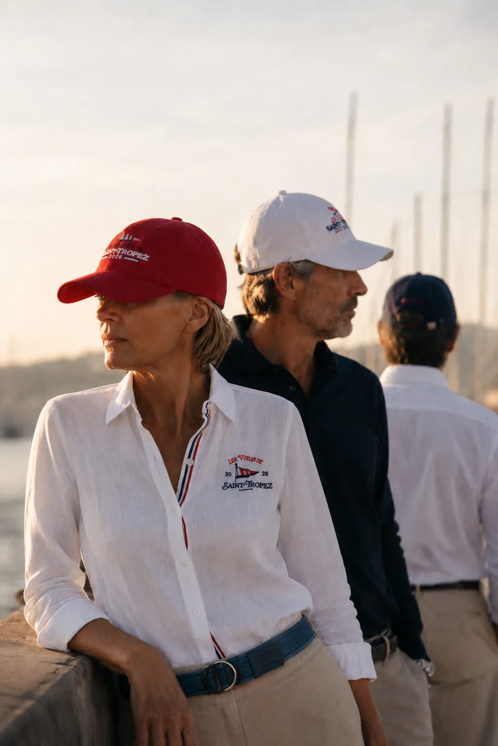 Trois personnes sur le quai au soleil couchant ; au premier plan, la casquette rouge de la collection 2026, une blanche et une marine derrière.