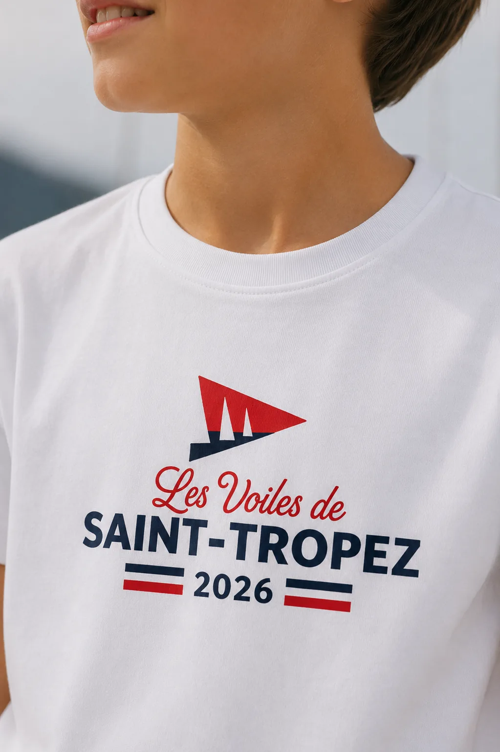 Macro du logo imprimé sur un t-shirt blanc enfant : pavillon rouge et marine, « Les Voiles de Saint-Tropez 2026 ».