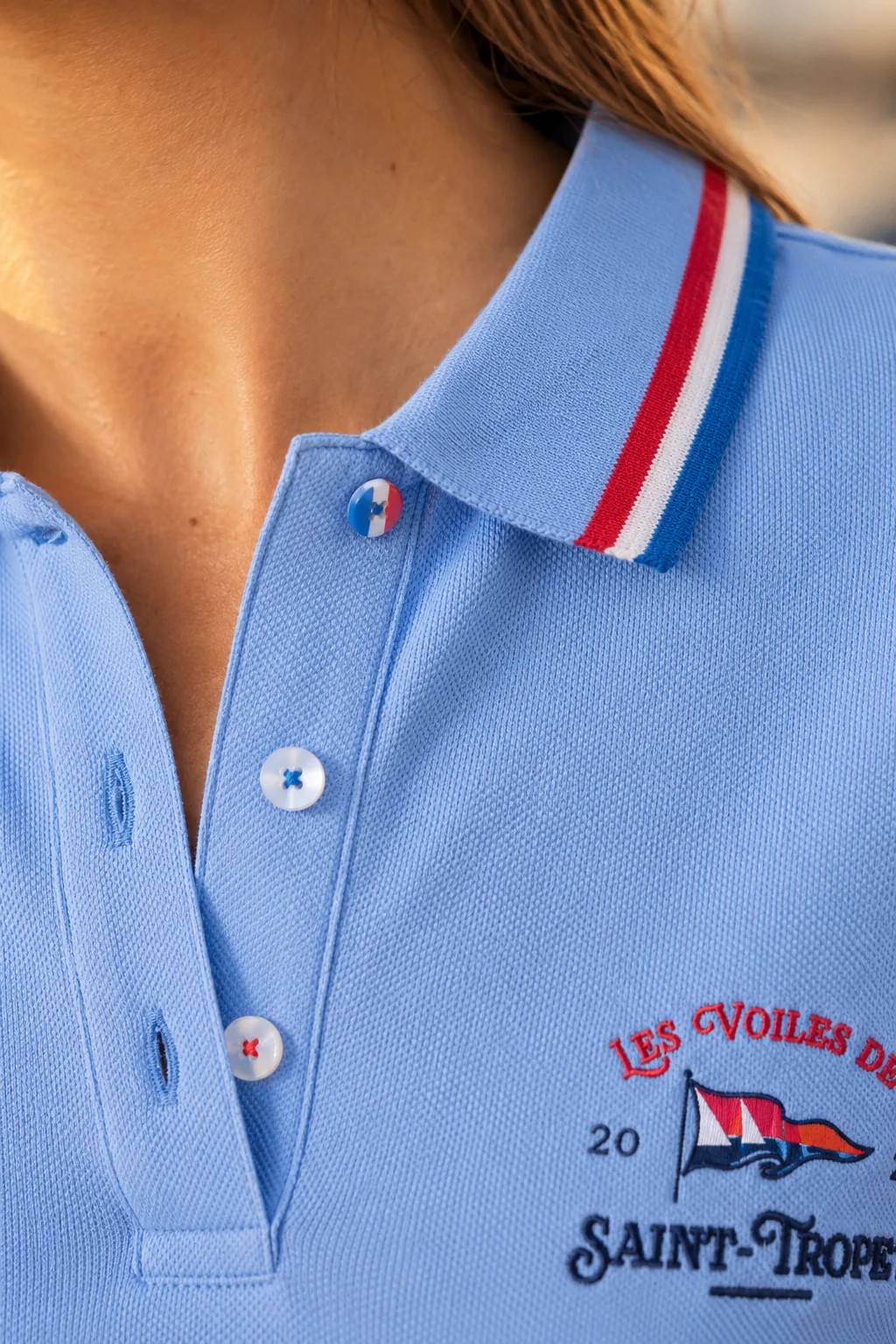 Macro du col et de la patte de boutonnage d'un polo femme bleu ciel : galon tricolore et boutons de nacre, dont un bleu-blanc-rouge.