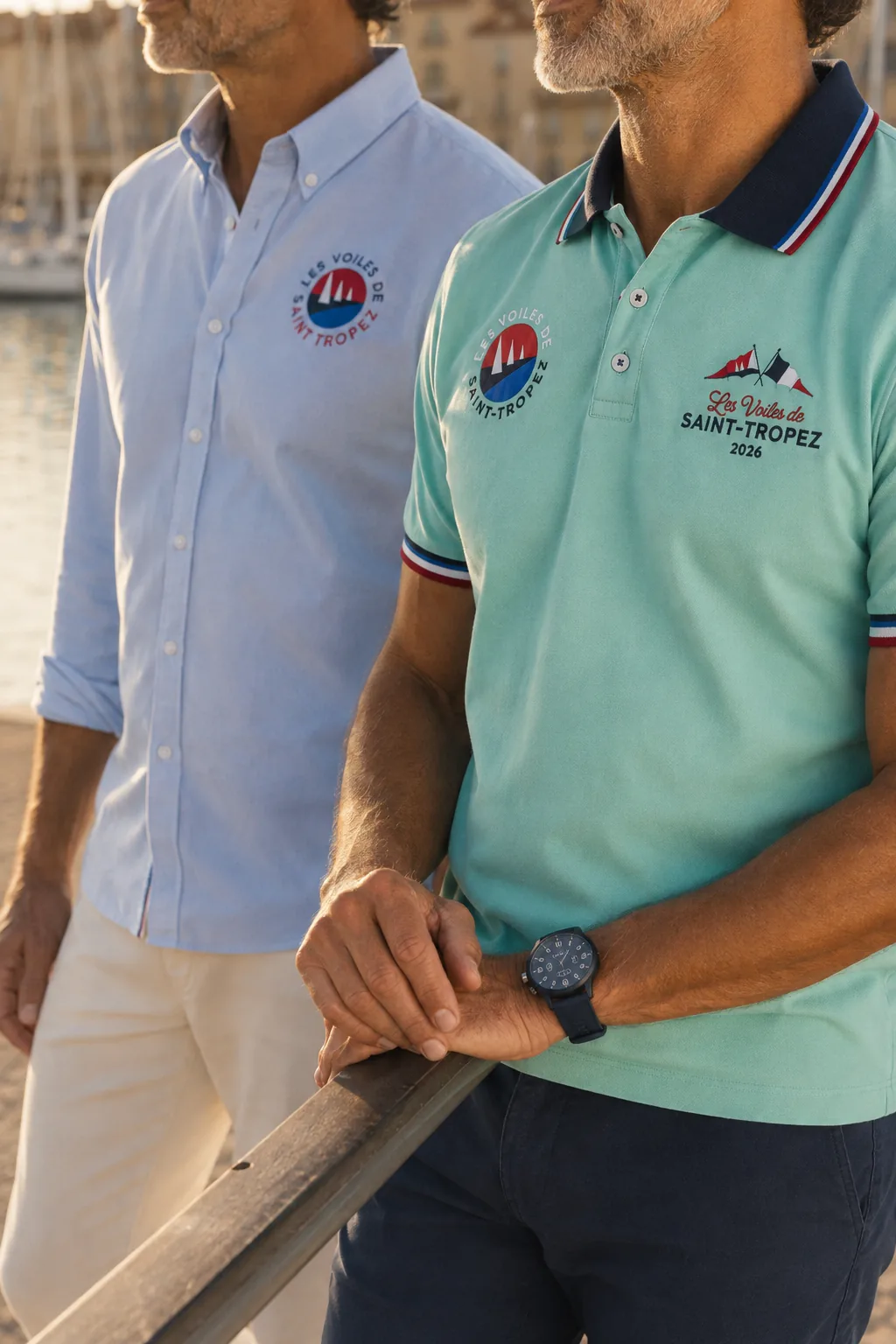 Plan rapproché de deux hommes au port : chemise Oxford bleu ciel et polo vert menthe, écusson « Les Voiles de Saint-Tropez 2026 » brodé sur la poitrine.