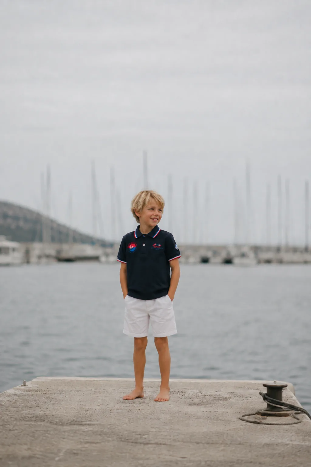 Un enfant en polo marine de la collection 2026, debout pieds nus au bout d'un ponton du port.