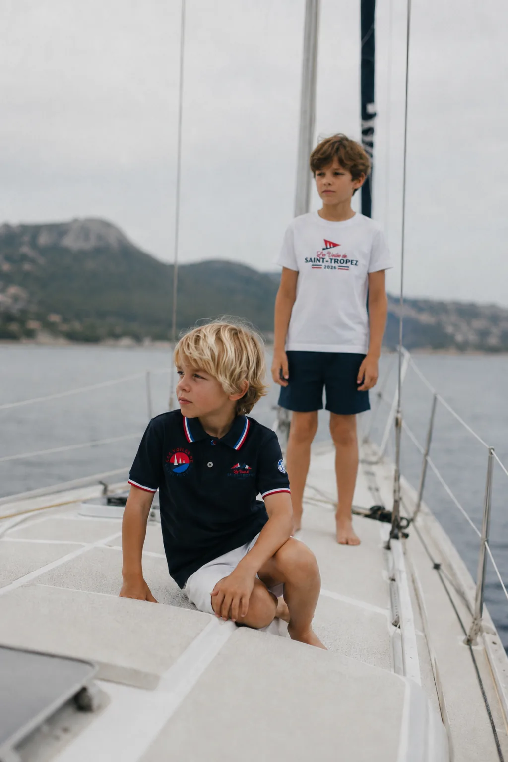 Deux enfants à bord d'un voilier, en polo marine et t-shirt blanc de la collection 2026.